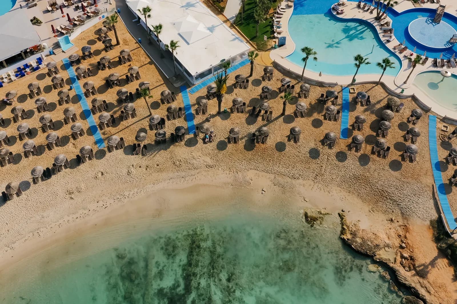 Vue aérienne d'un hôtel en bord de mer à Ayia Napa : piscines, transats et parasols de paille sur la plage, au bord d'une eau turquoise