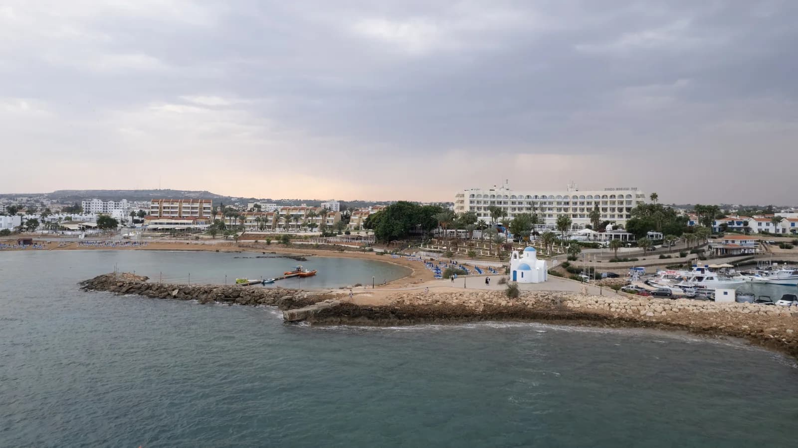 Front de mer d'Ayia Napa vu de la mer : jetée de pierres, petite chapelle blanche, port de plaisance et hôtels au fond