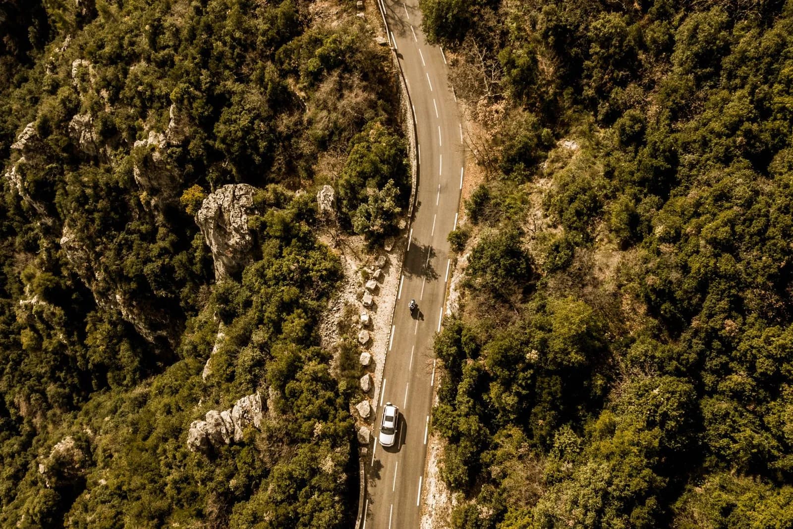 Vue aérienne d'une route sinueuse de montagne bordée de maquis méditerranéen, avec une voiture