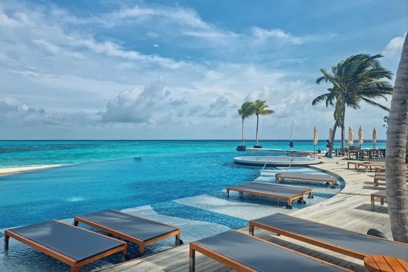 Piscine à débordement d'un resort balnéaire avec transats en bois et palmiers, face à la mer turquoise