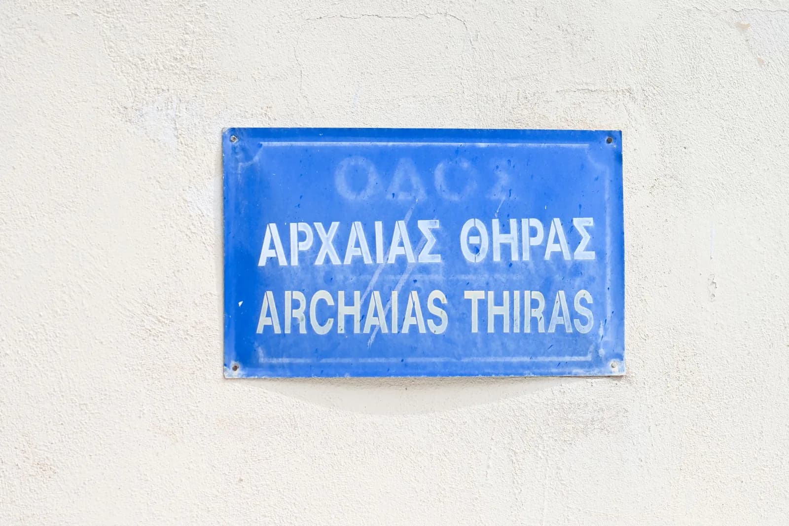 Plaque de rue bleue bilingue, en lettres grecques et en lettres latines, sur un mur blanc