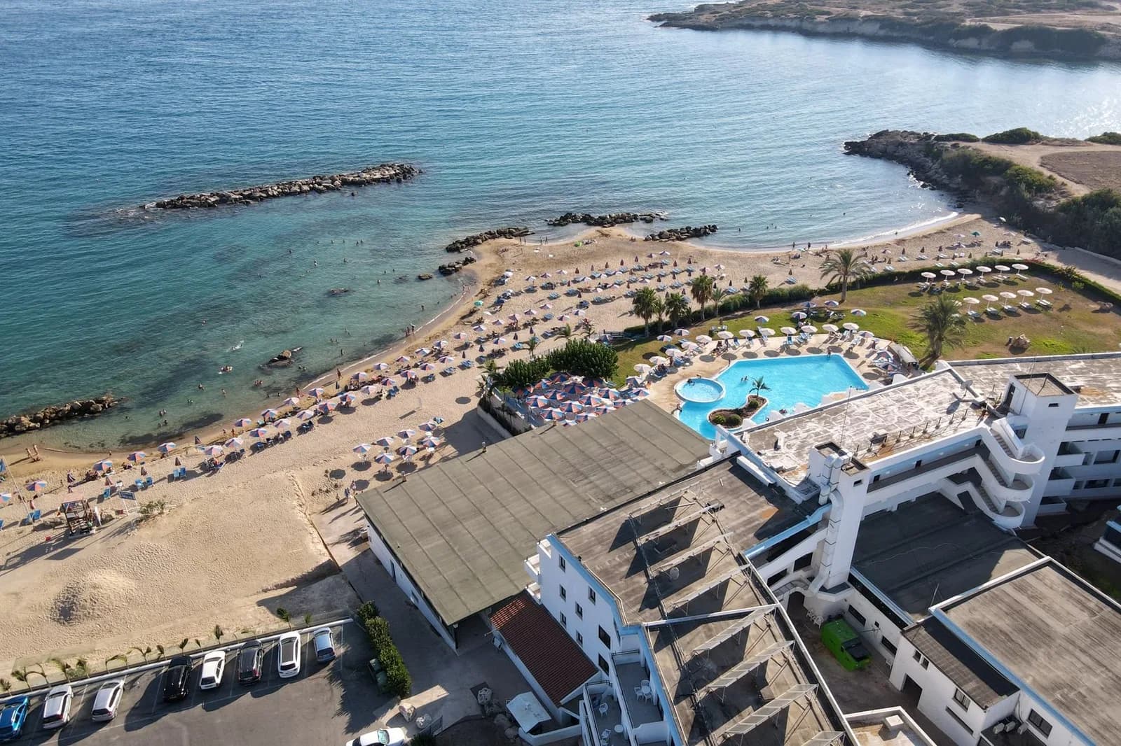 Vue aérienne d'un hôtel avec piscine face à une plage de sable aux parasols et à la mer turquoise, sur la côte de Paphos